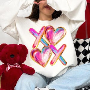 Puede incluir: Sudadera blanca con un dise&ntilde;o gr&aacute;fico de dos corazones rosas con contornos dorados y dos X doradas con pinceladas rosas, azules y amarillas.