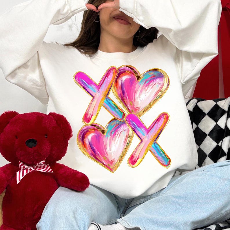 Puede incluir: Sudadera blanca con un dise&ntilde;o gr&aacute;fico de dos corazones rosas con contornos dorados y dos X doradas con pinceladas rosas, azules y amarillas.