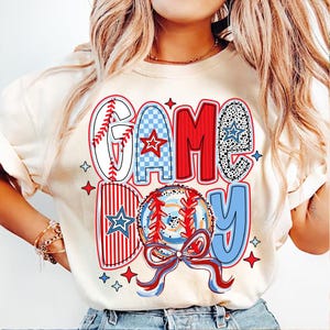 以下が含まれることがあります： 白いTシャツに、赤、白、青のグラフィックで「GAME DAY」と野球ボールと星が描かれています。