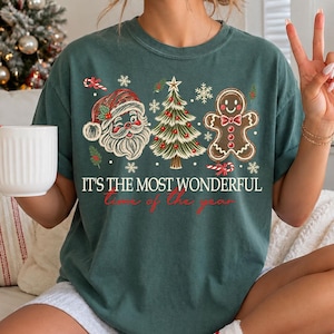 K&ouml;nnte beinhalten: Ein dunkelgr&uuml;nes T-Shirt mit einem Grafikdruck eines zwinkernden Weihnachtsmanns, eines geschm&uuml;ckten Weihnachtsbaums und eines Lebkuchenmanns. Der Text lautet IT'S THE MOST WONDERFUL time of the year, umgeben von kleinen wei&szlig;en Schneeflocken.