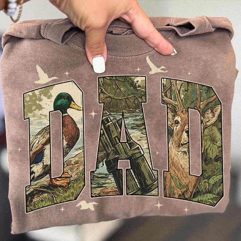 K&ouml;nnte beinhalten: Braunes T-Shirt mit dem Wort "DAD" in einem Design zum Thema Natur. Die Buchstaben zeigen eine Ente, ein Fernglas und ein Reh. Wei&szlig;e V&ouml;gel und Sterne sind um die Buchstaben herum gedruckt.