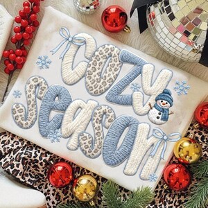 Pu&ograve; includere: Una maglietta a maniche lunghe color crema con le parole "COZY SEASON" in un design ricamato strutturato. Le lettere presentano un mix di motivi bianchi, blu e leopardati, con accenti di fiocchi di neve e una grafica di pupazzo di neve. Ornamenti natalizi circondano la maglietta.