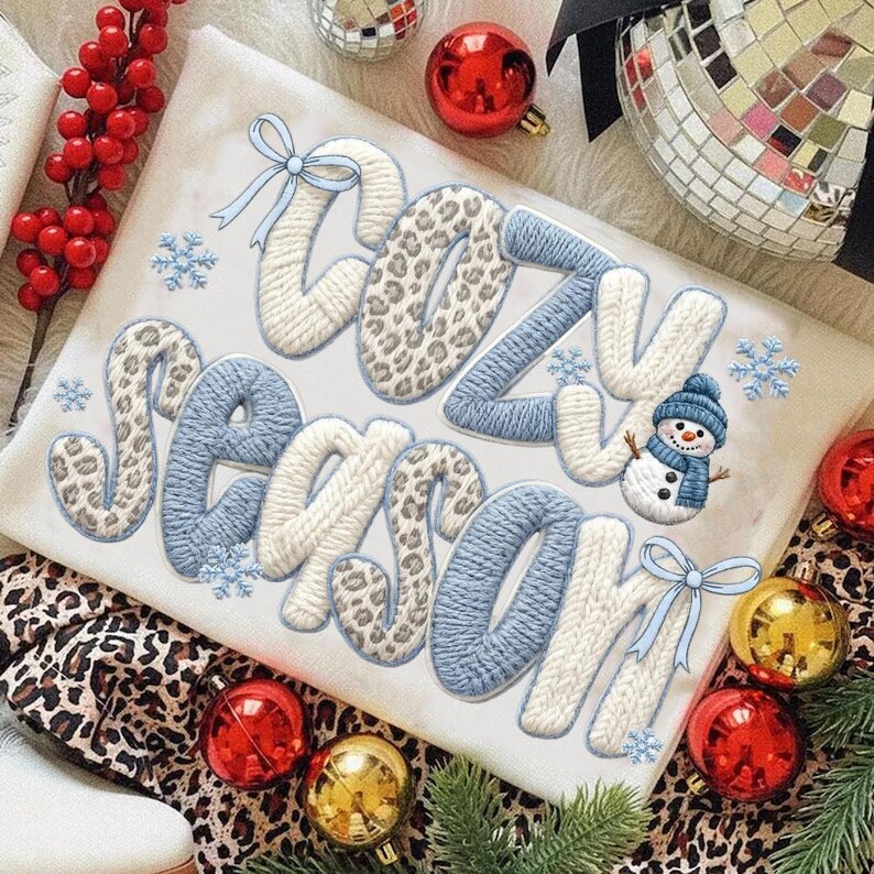 Pu&ograve; includere: Una maglietta a maniche lunghe color crema con le parole "COZY SEASON" in un design ricamato strutturato. Le lettere presentano un mix di motivi bianchi, blu e leopardati, con accenti di fiocchi di neve e una grafica di pupazzo di neve. Ornamenti natalizi circondano la maglietta.