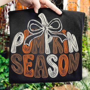 May include: A black t-shirt featuring a graphic design of a pumpkin outline topped with a large bow. The text "PUMPKIN SEASON" is written in a stylized, scribbled font in shades of orange, brown, and white, centered on the front of the shirt.