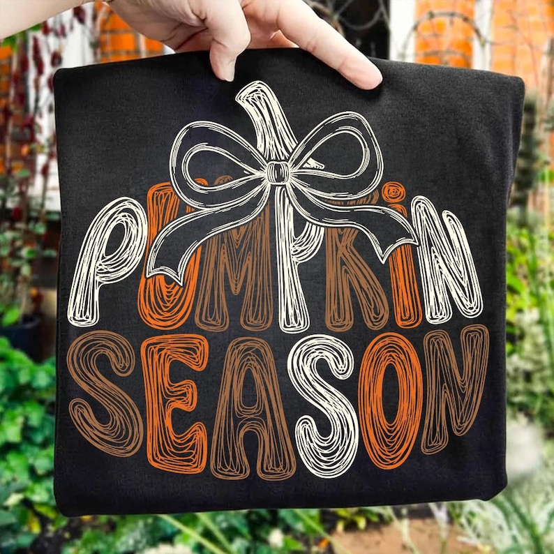 May include: A black t-shirt featuring a graphic design of a pumpkin outline topped with a large bow. The text "PUMPKIN SEASON" is written in a stylized, scribbled font in shades of orange, brown, and white, centered on the front of the shirt.