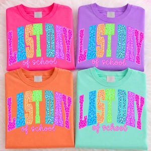 May include: Four colorful t-shirts in pink, purple, orange, and teal. Each shirt features the text "LAST DAY of school" in a bold, multi-colored, leopard print design. The shirts are neatly folded and displayed on a white, fluffy surface.