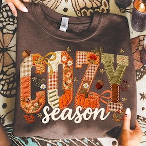 May include: A dark brown crewneck sweatshirt featuring the text COZY season in a patchwork-style font. The letters are decorated with autumn-themed patterns including orange pumpkins, brown and white gingham, floral prints, and yellow bows.