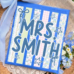 Könnte beinhalten: Blau-weißes Dekoschild mit dem Text "MRS SMITH" in großen, blau gepunkteten Buchstaben. Das Schild hat einen gestreiften Hintergrund mit Blumen- und Vichy-Mustern, akzentuiert mit Sternformen und Schleifen.