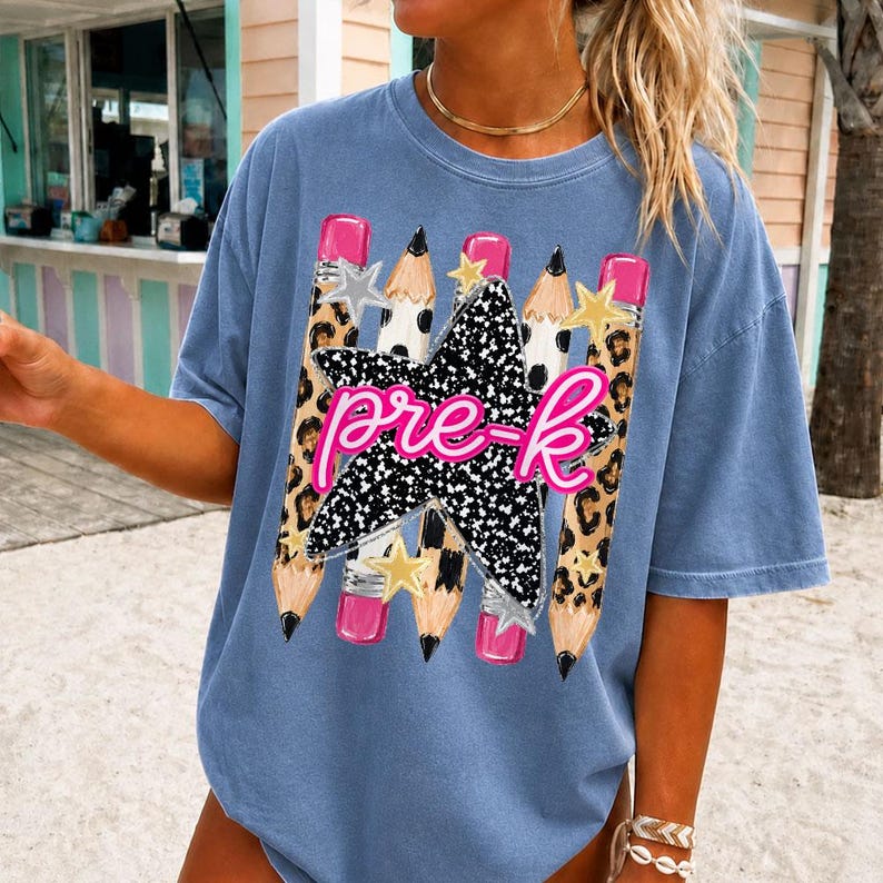May include: A blue t-shirt featuring a graphic print of three pencils with leopard print patterns and pink erasers. A large black star with white speckles is centered over the pencils, overlaid with the pink cursive text pre-k.