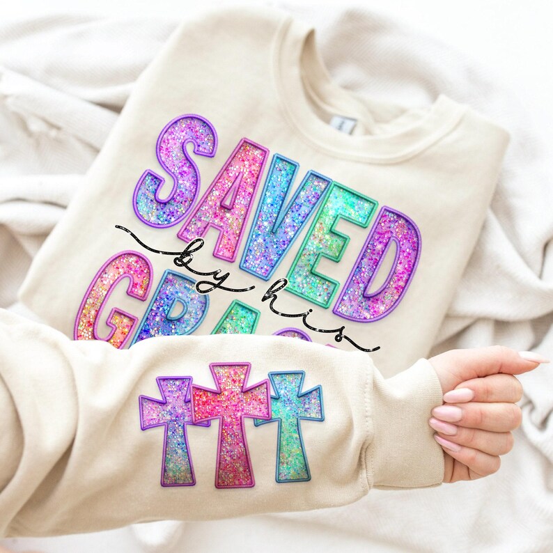 May include: Beige sweatshirt with the words "SAVED BY GRACE" in colorful glitter lettering. The sleeve features three glitter cross designs in purple, pink, and blue. The sweatshirt is folded on a white surface.