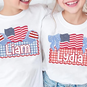 Puede incluir: Camisetas blancas con dise&ntilde;os patri&oacute;ticos. Una camiseta presenta el nombre "Liam" con un tema rojo, blanco y azul, incluyendo una gorra de b&eacute;isbol y la bandera americana. La otra camiseta tiene "Lydia" con lazos y banderas.