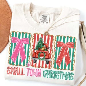 Könnte beinhalten: Cremefarbenes T-Shirt mit Weihnachtsgrafik. Das Design zeigt einen roten Truck mit einem Weihnachtsbaum, rosa und rote Schleifen und den Text "SMALL TOWN CHRISTMAS". Eine goldene Halskette liegt auf dem Shirt.