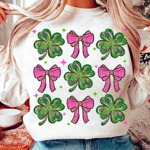 May include: White sweatshirt featuring a pattern of green and pink shamrocks and bows. The shamrocks have green and pink glitter accents, and the bows are a bright pink. Green and pink stars are scattered around the design.