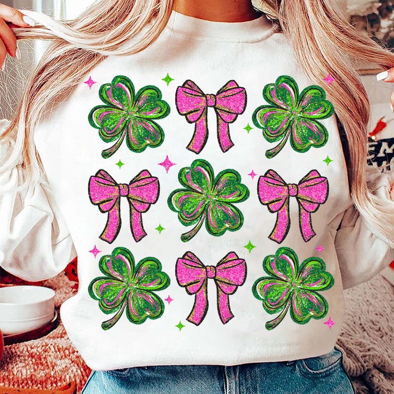 May include: White sweatshirt featuring a pattern of green and pink shamrocks and bows. The shamrocks have green and pink glitter accents, and the bows are a bright pink. Green and pink stars are scattered around the design.