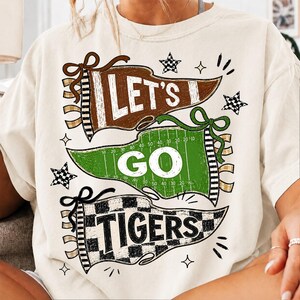 May include: A cream-colored t-shirt featuring a graphic design of three pennants. The top brown pennant says LET'S, the middle green football field pennant says GO, and the bottom checkered pennant says TIGERS. The design includes small stars.