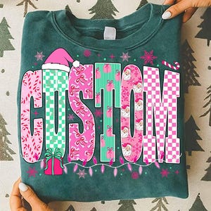 May include: A folded teal sweatshirt featuring the word "CUSTOM" in a festive design. The letters are decorated with pink bows, Santa faces, and a pink and white checkerboard pattern. A Santa hat and string lights add to the holiday theme.