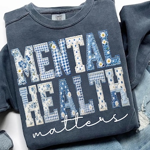 May include: A blue sweatshirt with the words "MENTAL HEALTH matters" in a patchwork design. The letters are made of various blue fabrics, including floral, gingham, and polka dot patterns. The sweatshirt has a crew neck and long sleeves.