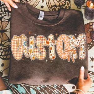 Puede incluir: Una sudadera marrón con la palabra "CUSTOM" en una fuente decorativa. Las letras están rellenas de varios patrones, incluyendo cuadros, flores y estampado de leopardo, y están delineadas con un borde festoneado. La sudadera es sostenida por una persona.