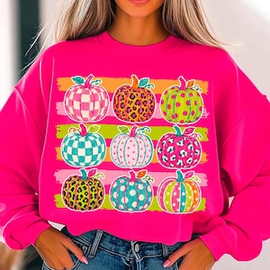 May include: A bright hot pink long-sleeve sweatshirt featuring a graphic print of nine pumpkins arranged in a three-by-three grid. Each pumpkin displays a unique pattern, including leopard print, polka dots, and checkered designs in vibrant colors.
