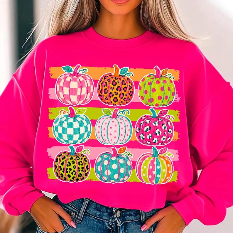May include: A bright hot pink long-sleeve sweatshirt featuring a graphic print of nine pumpkins arranged in a three-by-three grid. Each pumpkin displays a unique pattern, including leopard print, polka dots, and checkered designs in vibrant colors.