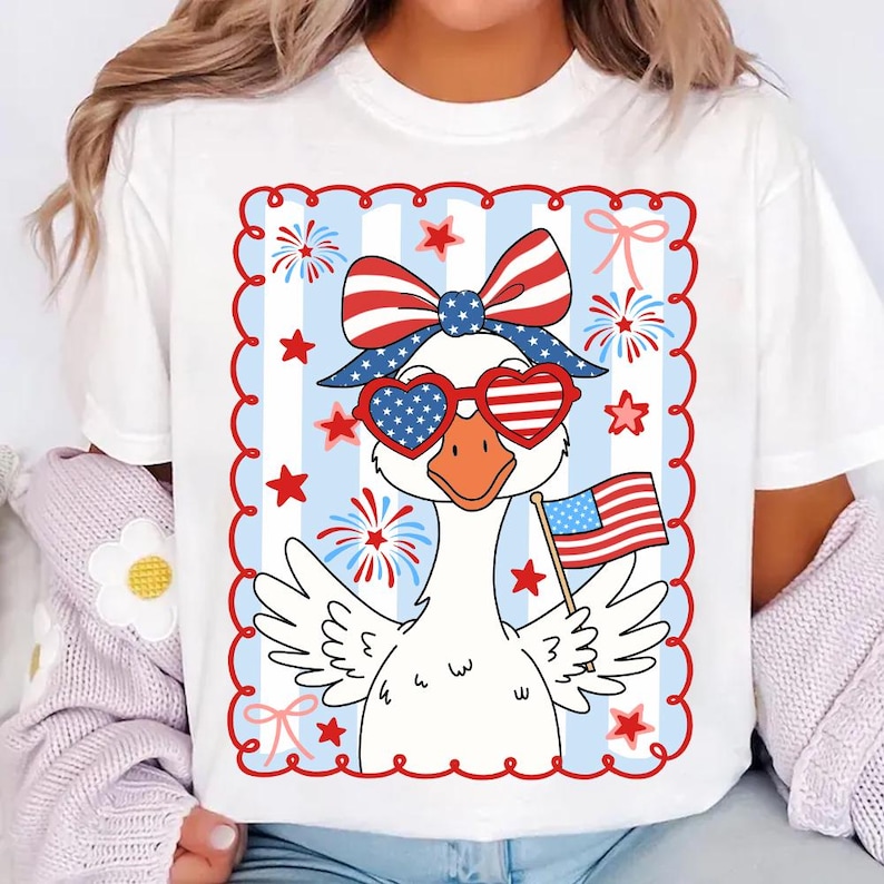 Puede incluir: Camiseta blanca con un dibujo de un ganso con gafas de sol en forma de coraz&oacute;n y un lazo patri&oacute;tico. El ganso sostiene una peque&ntilde;a bandera americana. El dise&ntilde;o incluye elementos rojos, blancos y azules, estrellas y fuegos artificiales.