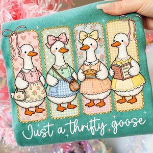Puede incluir: Camiseta de color turquesa con cuatro gansos de dibujos animados con vestidos y lazos, cada uno con un atuendo diferente. Los gansos están en marcos rectangulares con fondos florales y de lunares. El texto "Just a thrifty goose" está escrito en blanco.