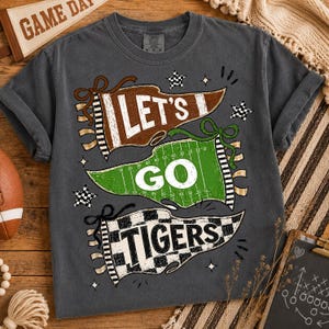 May include: A charcoal gray short-sleeve t-shirt featuring a graphic print of three pennants. The top pennant is brown with the text LET'S, the middle is green with white yard lines and the text GO, and the bottom is black and white checkered with TIGERS.