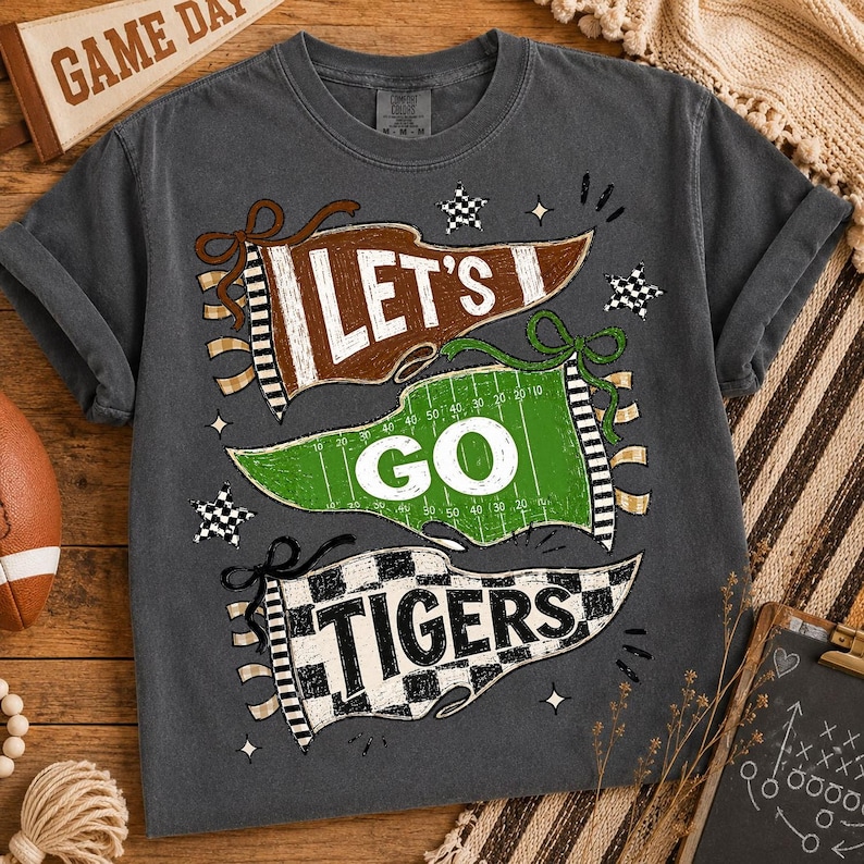 May include: A charcoal gray short-sleeve t-shirt featuring a graphic print of three pennants. The top pennant is brown with the text LET'S, the middle is green with white yard lines and the text GO, and the bottom is black and white checkered with TIGERS.