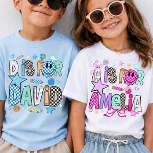 May include: A light blue t-shirt with the text D IS FOR DAVID and a white t-shirt with the text A IS FOR AMELIA. Both shirts feature colorful school-themed graphics including checkered patterns, smiley faces, stars, pencils, and school buses.