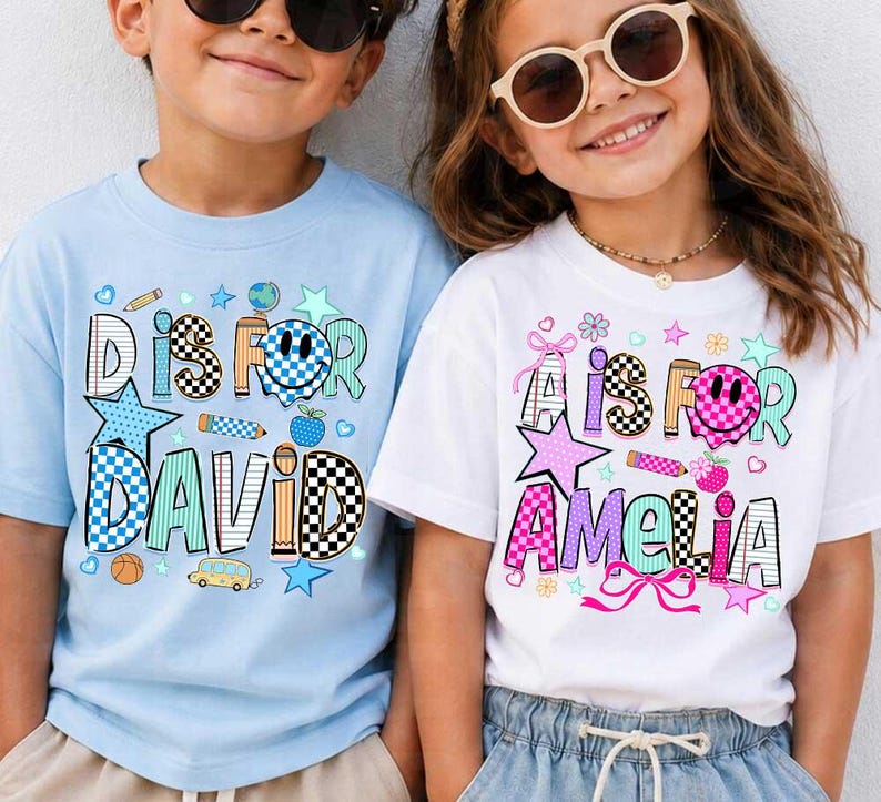 May include: A light blue t-shirt with the text D IS FOR DAVID and a white t-shirt with the text A IS FOR AMELIA. Both shirts feature colorful school-themed graphics including checkered patterns, smiley faces, stars, pencils, and school buses.