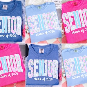 May include: A collection of six t-shirts in various colors: blue, light blue, and pink. Each shirt features the word "SENIOR" in large, sequined letters, with "class of 2026" below. The shirts are casual and likely for graduation.
