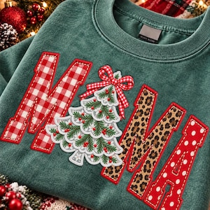 Op de afbeelding: Een donkergroene sweater met ronde hals en het woord MAMA in grote letters. De M is rood-wit geblokt, de A is een kerstboom van kant met een rood-wit geblokte strik, de M heeft een luipaardprint en de A is rood met witte stippen.