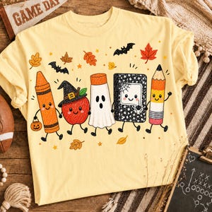May include: A pale yellow t-shirt featuring a colorful graphic of five anthropomorphic school supplies: an orange crayon holding a pumpkin, a red apple wearing a witch hat, a white ghost, a black notebook with a spider, and a yellow pencil.
