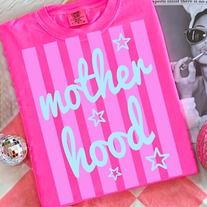 Puede incluir: Camiseta rosa brillante con rayas verticales rosas y la palabra "motherhood" en escritura azul claro. Estrellas azules claras est&aacute;n dispersas alrededor del texto. La camiseta es de la marca Comfort Colors.