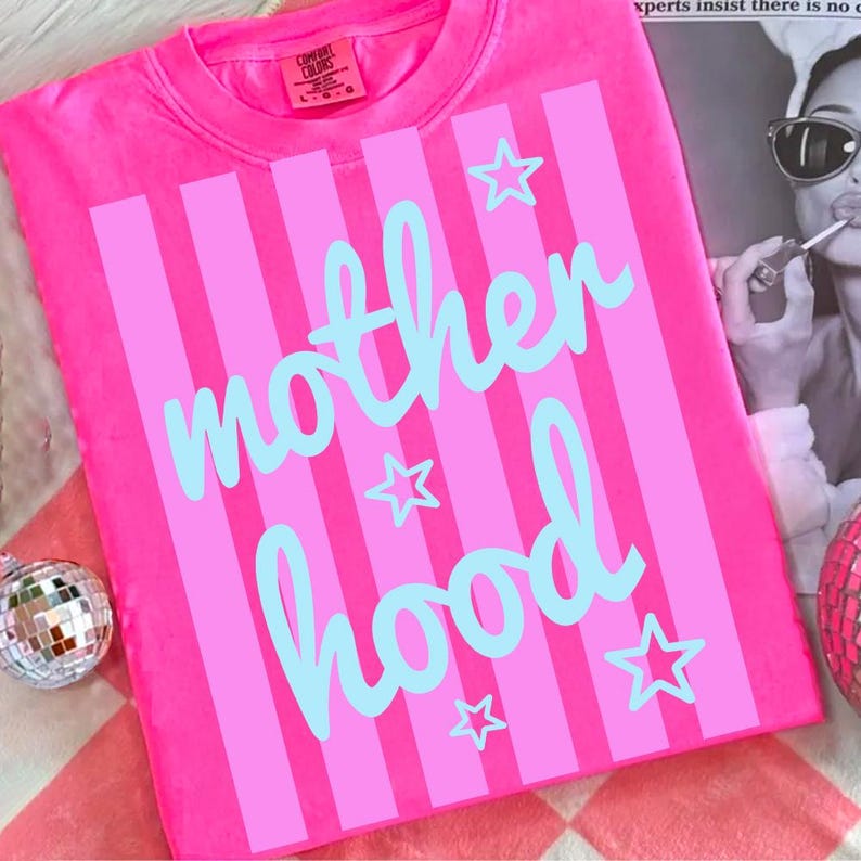 Puede incluir: Camiseta rosa brillante con rayas verticales rosas y la palabra "motherhood" en escritura azul claro. Estrellas azules claras est&aacute;n dispersas alrededor del texto. La camiseta es de la marca Comfort Colors.