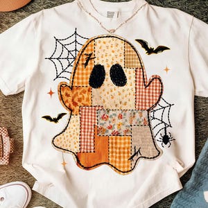 May include: A white short-sleeve t-shirt featuring a large central graphic of a ghost made of various orange, yellow, and brown patchwork patterns. The design includes black spiderwebs, two flying bats, a small spider, and orange star accents.