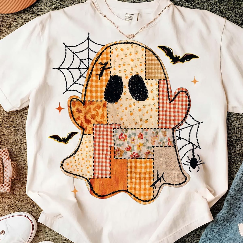 May include: A white short-sleeve t-shirt featuring a large central graphic of a ghost made of various orange, yellow, and brown patchwork patterns. The design includes black spiderwebs, two flying bats, a small spider, and orange star accents.
