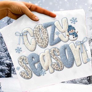 Pu&ograve; includere: Un tessuto bianco con le parole "COZY SEASON" ricamate in blu, bianco e stampa leopardata. &Egrave; presente un piccolo pupazzo di neve con cappello e sciarpa blu. Fiocchi di neve e fiocchi azzurri completano il tema invernale.