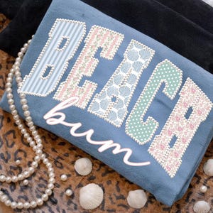 Puede incluir: Una camiseta azul con las palabras "BEACH bum" en una fuente decorativa. Las letras están estampadas con diseños de rayas, flores y conchas marinas. Un collar de perlas y conchas marinas están dispuestas alrededor de la camiseta.