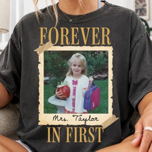 May include: A charcoal gray t-shirt featuring a vintage-style photo print of a young child holding a red apple and a pink lunch bag. The text FOREVER is printed above the photo, and Mrs. Taylor IN FIRST is printed below the photo in gold lettering.
