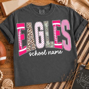 May include: A charcoal gray short-sleeve t-shirt featuring the word EAGLES in large, multi-patterned letters including leopard print, pink polka dots, camouflage, and pink stripes, with the text school name printed in white underneath the word.