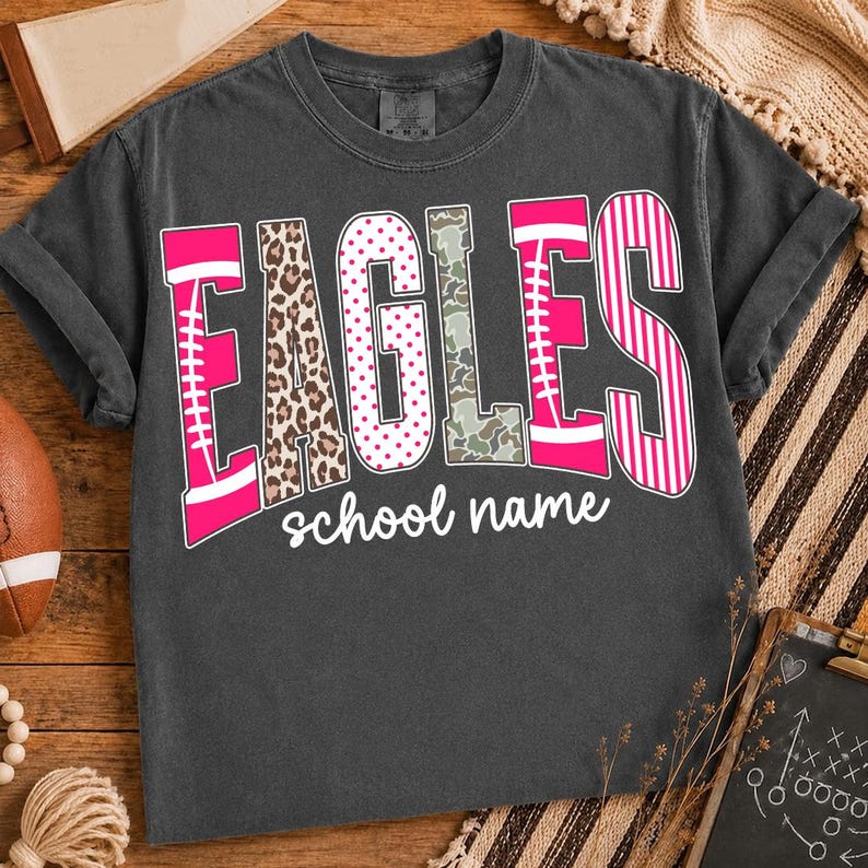 May include: A charcoal gray short-sleeve t-shirt featuring the word EAGLES in large, multi-patterned letters including leopard print, pink polka dots, camouflage, and pink stripes, with the text school name printed in white underneath the word.