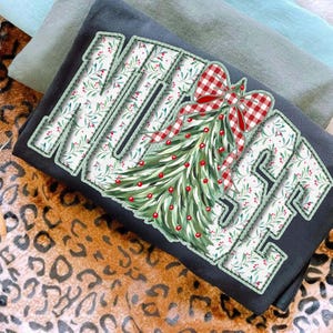 Op de afbeelding: Een zwart sweatshirt met het woord "MERRY" in een wit bloemenpatroon met groene randen. Een kerstboomontwerp met rode bessen en een rood-witte geruite strik staat rechts. Twee andere sweatshirts zijn op de achtergrond te zien.