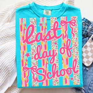 Puede incluir: Una camiseta turquesa con el texto "Last day of School" en rosa. El dise&ntilde;o presenta rayas verticales con patrones florales y de estrellas, y lazos rosas. La camiseta se muestra sobre una superficie blanca con vaqueros y una funda de tel&eacute;fono en el fondo.