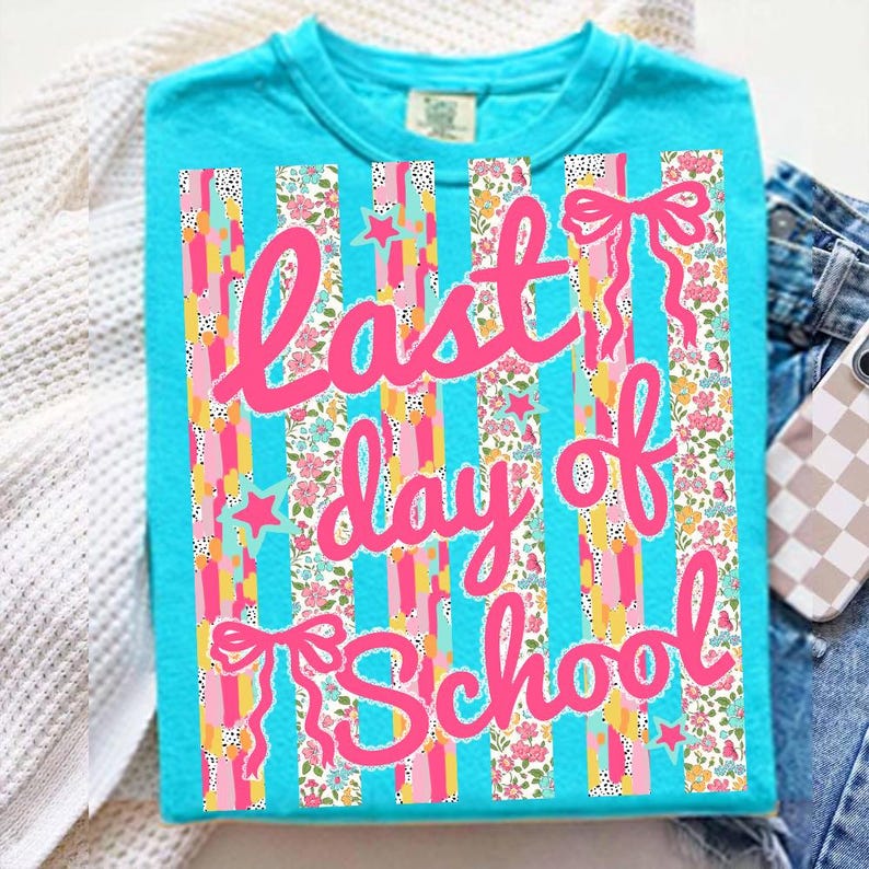 Puede incluir: Una camiseta turquesa con el texto "Last day of School" en rosa. El dise&ntilde;o presenta rayas verticales con patrones florales y de estrellas, y lazos rosas. La camiseta se muestra sobre una superficie blanca con vaqueros y una funda de tel&eacute;fono en el fondo.