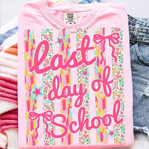 Puede incluir: Camiseta rosa claro con la frase "Last Day of School" en rosa brillante. El dise&ntilde;o incluye rayas verticales de varios colores y estampados florales, con lazos rosas y estrellas. La camiseta est&aacute; doblada.