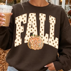 May include: A dark brown crewneck sweatshirt featuring the word FALL in large, cream-colored letters with brown polka dots. Below the text is a graphic of a pumpkin with a plaid and floral pattern, accented by a light pink bow on its stem.