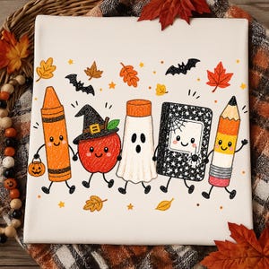May include: A white t-shirt featuring a colorful graphic of five anthropomorphic school supplies and a ghost holding hands. The design includes an orange crayon, a red apple wearing a witch hat, a white ghost, a black notebook, and a yellow pencil.