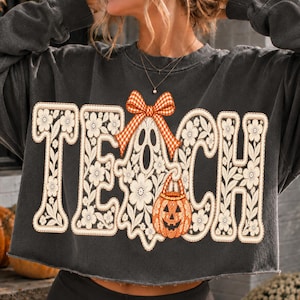 May include: A charcoal grey long-sleeve cropped sweatshirt featuring the word TEACH in large, cream-coloured floral lace-style letters. A ghost graphic with an orange and white checkered bow and a jack-o'-lantern bucket is centred.