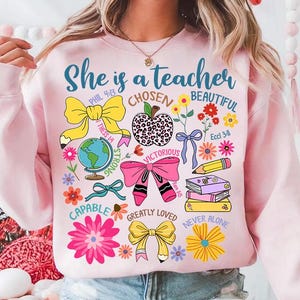 以下が含まれることがあります： 「She is a teacher」という文字と、リボン、花、本、地球儀のデザインが施されたピンク色のスウェットシャツ。デザインには、聖書の言葉や「Chosen」、「Beautiful」、「Victorious」、「Capable」、「Greatly Loved」、「Never Alone」などの言葉も含まれています。